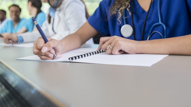 An unrecognizable medical student sits at a table in her lecture hall and writes in her class workbook.
